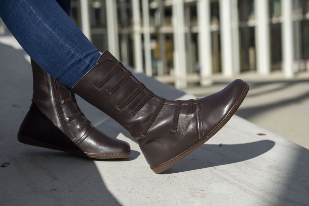 En person i ZAQQs ATTRAQT Stretch Nappa Brown-stövlar och blå jeans sitter på en betongyta. I bakgrunden syns en modern glasfasad av en byggnad.