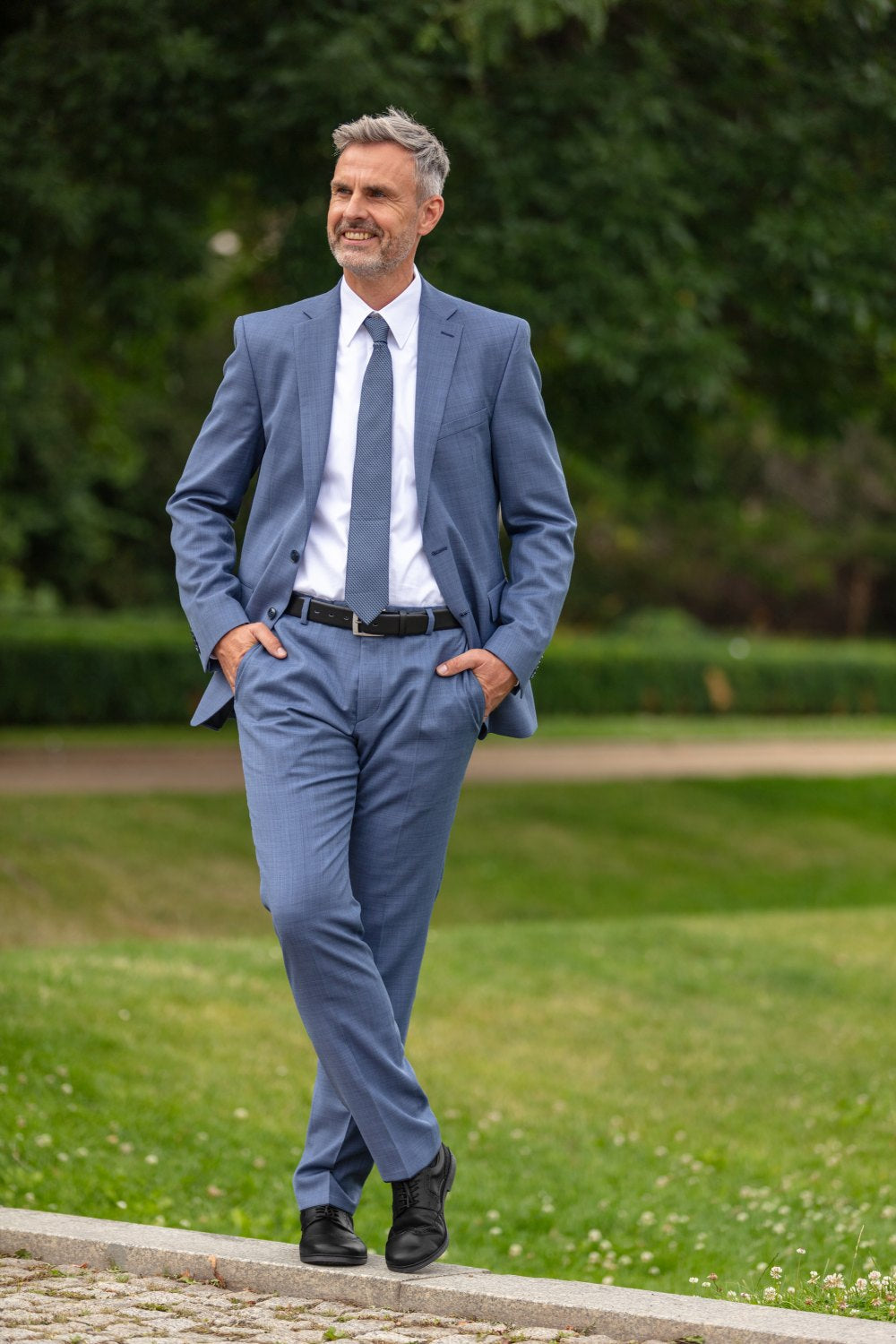 En man i blå kostym och slips går med händerna i fickorna utomhus på en stig. Han ler, har grått hår och skägg och står framför en bakgrund av grönt gräs och träd. Han bär BRIQ Brogue Black från ZAQQ, som kompletterar hans eleganta look.