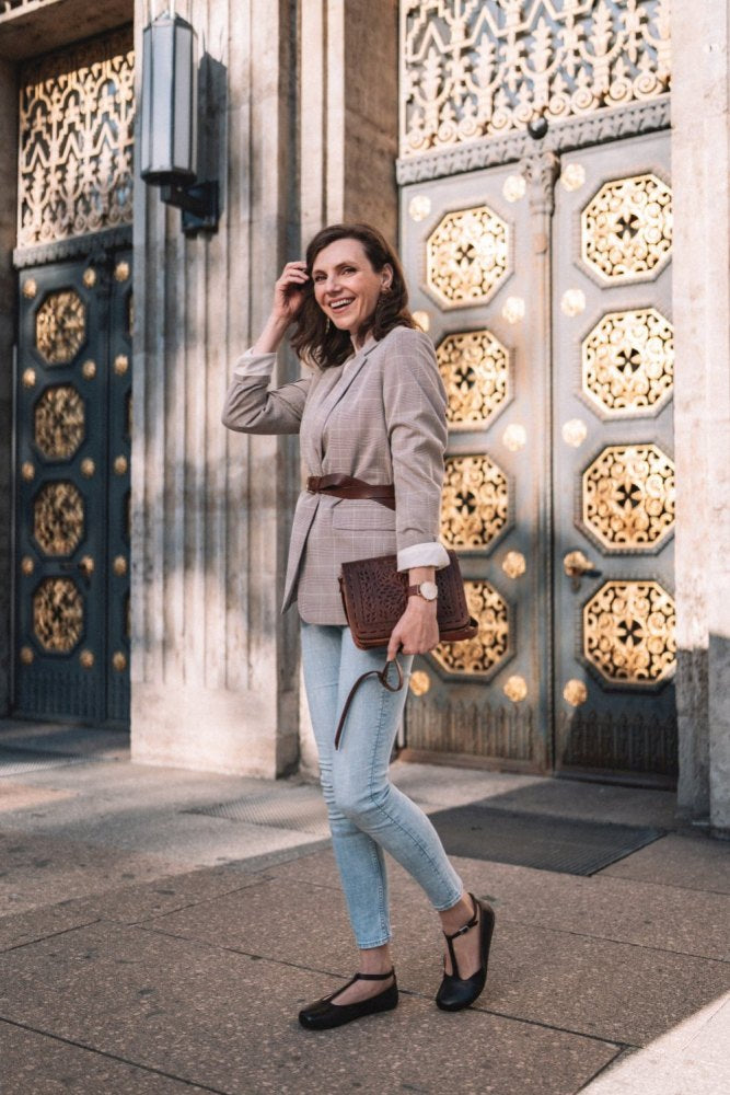 En person i rutig blazer och jeans står på gatan och håller en strukturerad clutch i handen. I bakgrunden syns dörrar med invecklat guldmönster. Hon bär en klocka på handleden och eleganta svarta skor av slätt läder från ZAQQ, kända som modellen QITE Dark Brown.