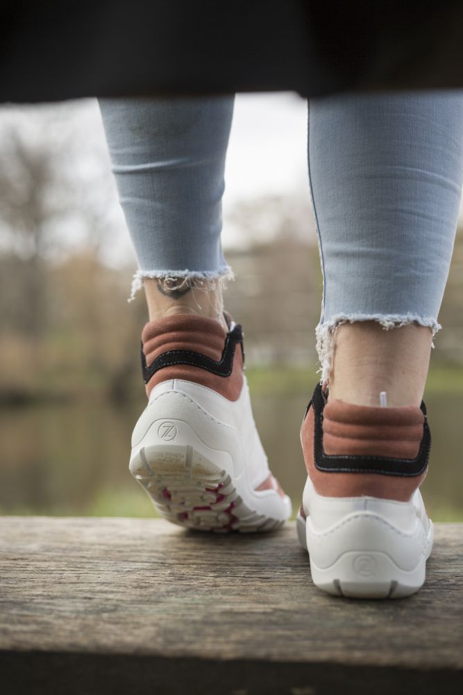 En person i fransiga, ljusblå jeans och vita sneakers med rosa och svarta accenter står på en träyta. De ZAQQ VAQATION Rosa Waterproof-skorna erbjuder en oöverträffad andningsförmåga. I bakgrunden syns en suddig vy över en park med träd och en damm.